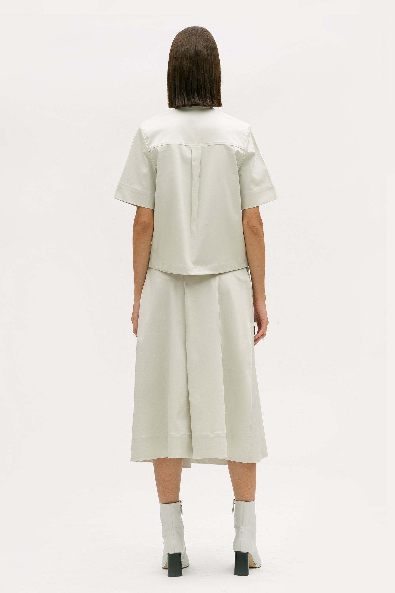 A woman with shoulder length brown hair is wearing an off-white utility shirt, a long skirt and white boots
