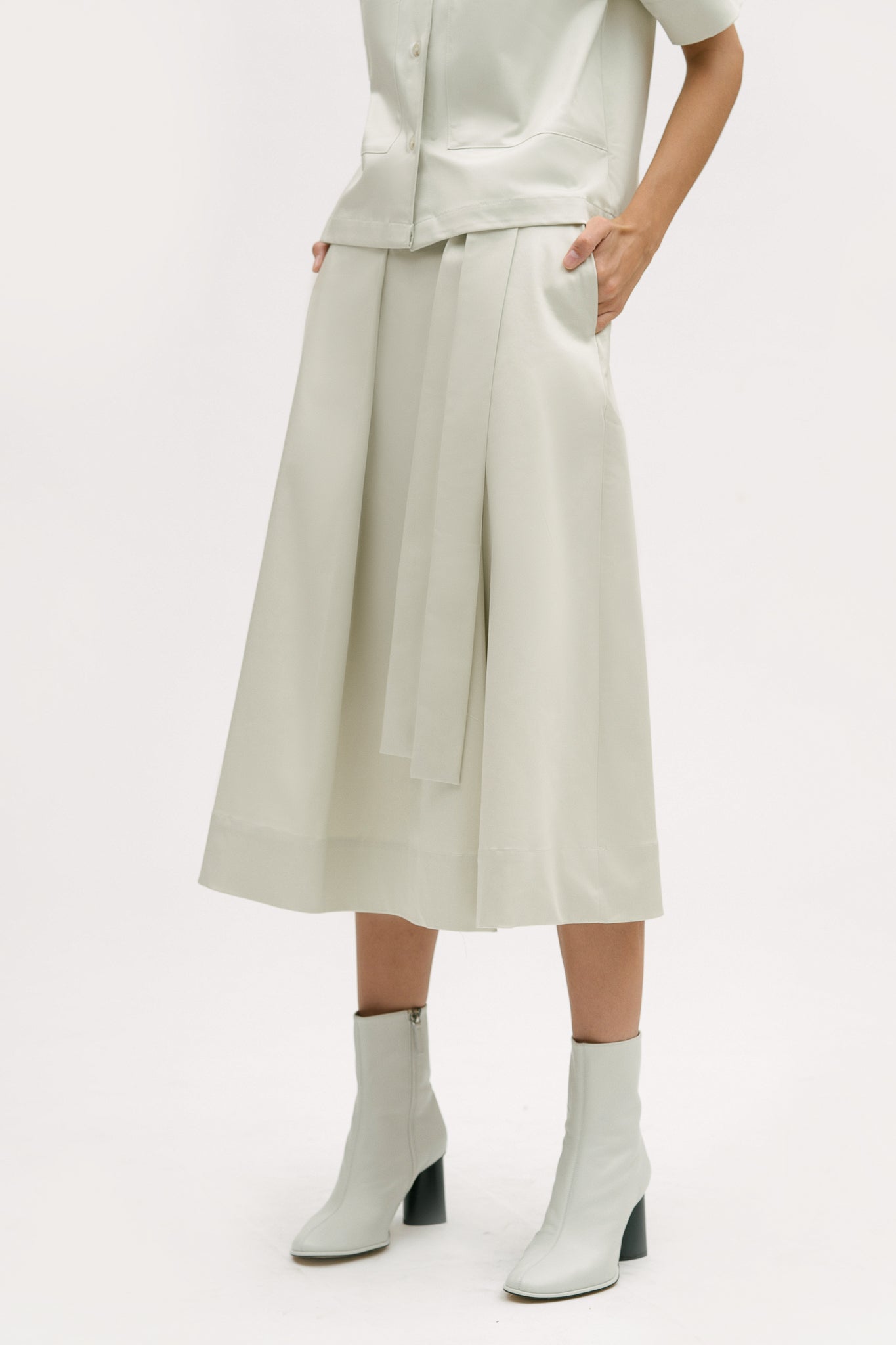 A woman with shoulder length brown hair is wearing an off-white shirt and calf length skirt. She is also wearing white boots.