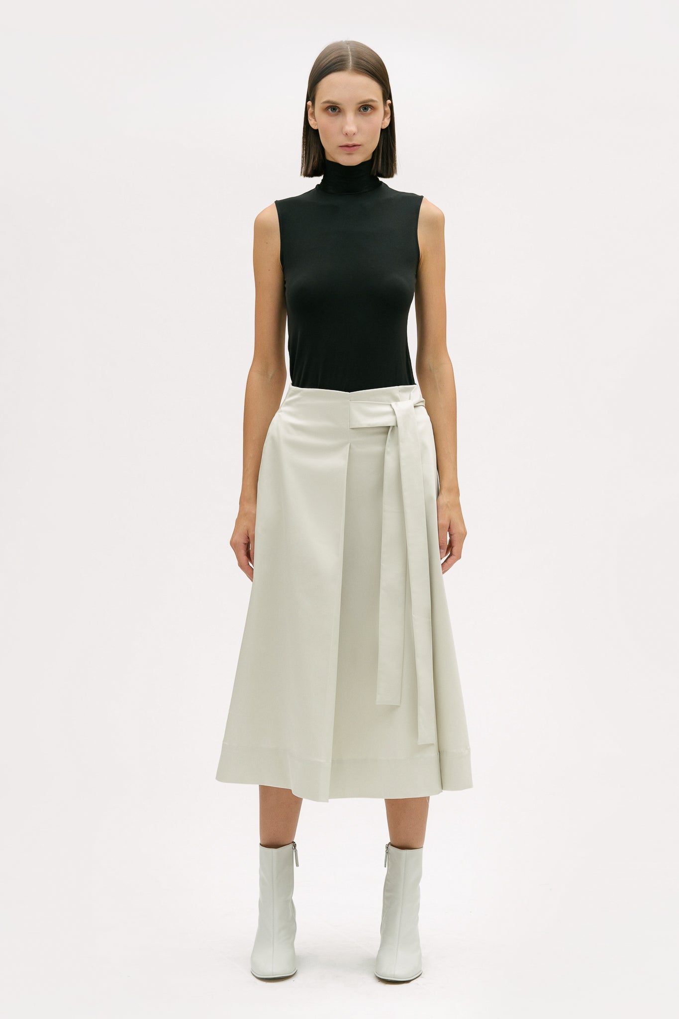 A woman with shoulder length brown hair is wearing a black top and off-white calf length skirt. She is also wearing white boots.