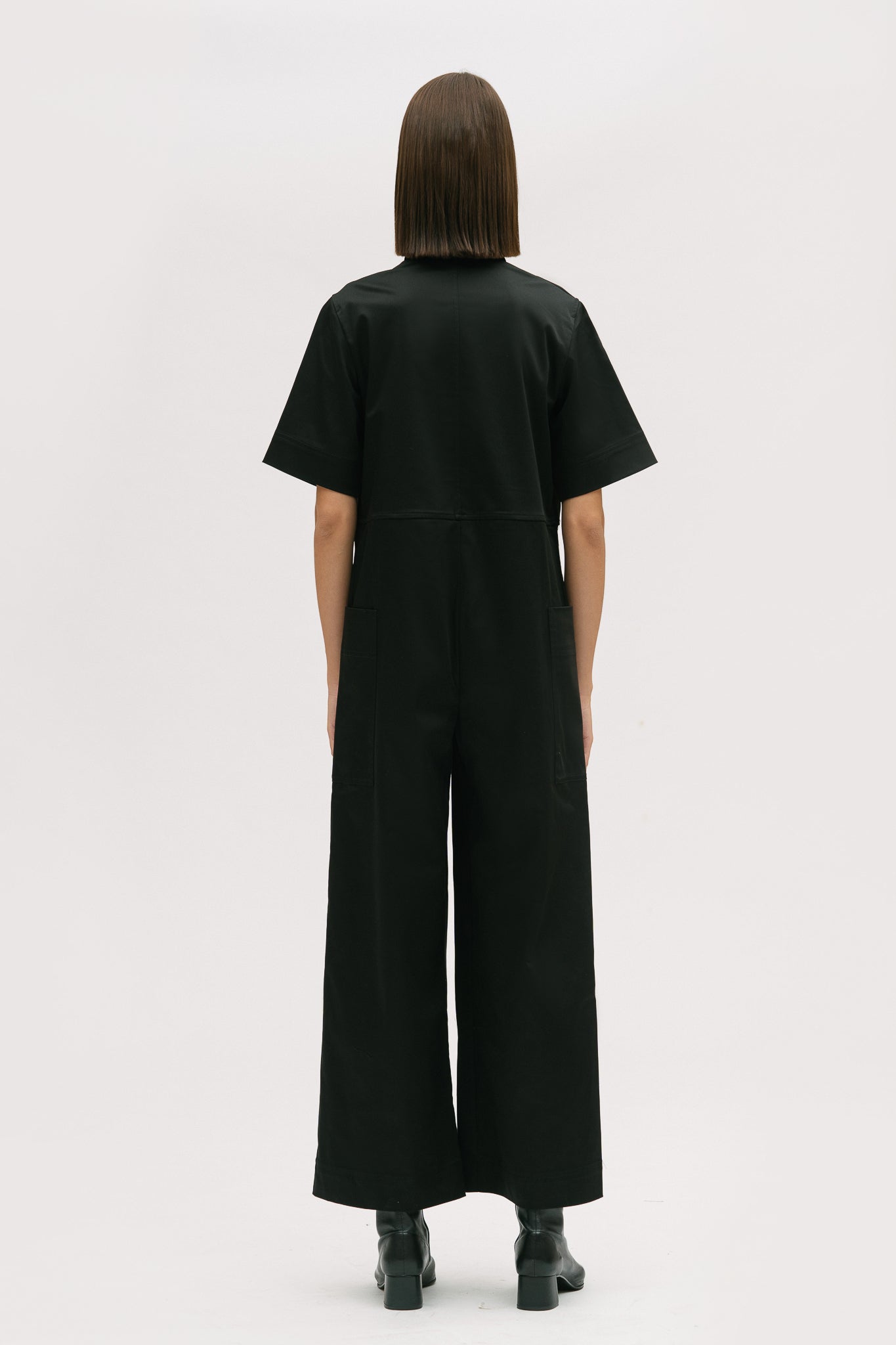 A woman with short brown hair is wearing a black collared jumpsuit with a tie belt. She is wearing black boots.