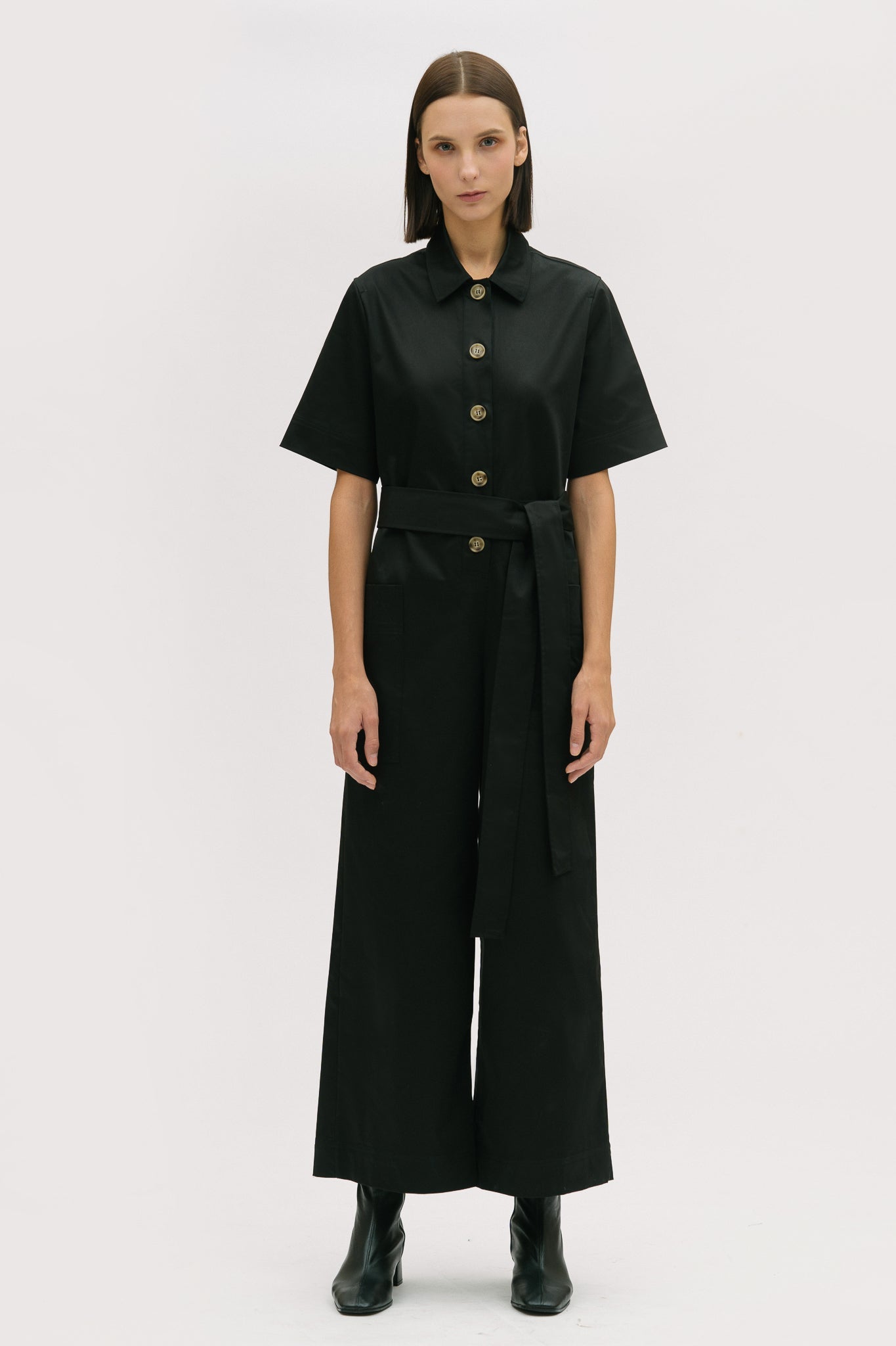 A woman with short brown hair is wearing a black collared jumpsuit with a tie belt. She is wearing black boots.