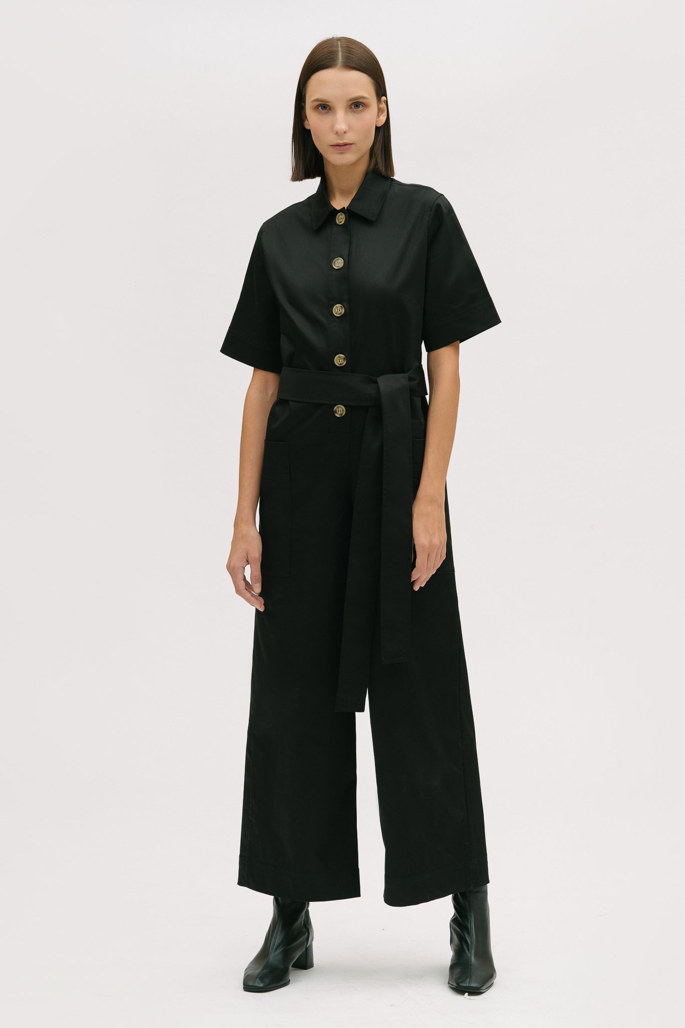 A woman with short brown hair is wearing a black collared jumpsuit with a tie belt. She is wearing black boots.
