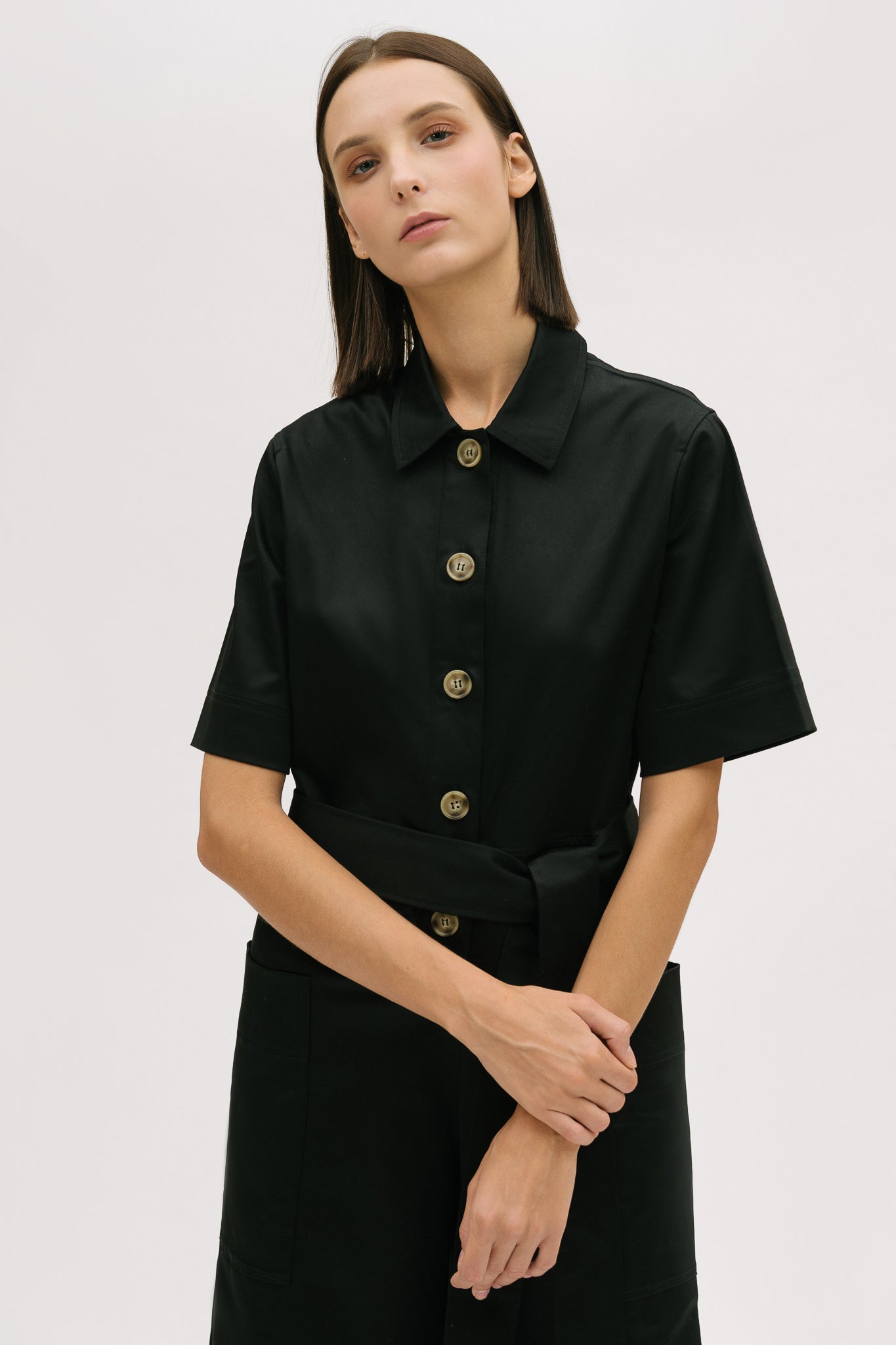 A woman with short brown hair is wearing a black collared jumpsuit with a tie belt.