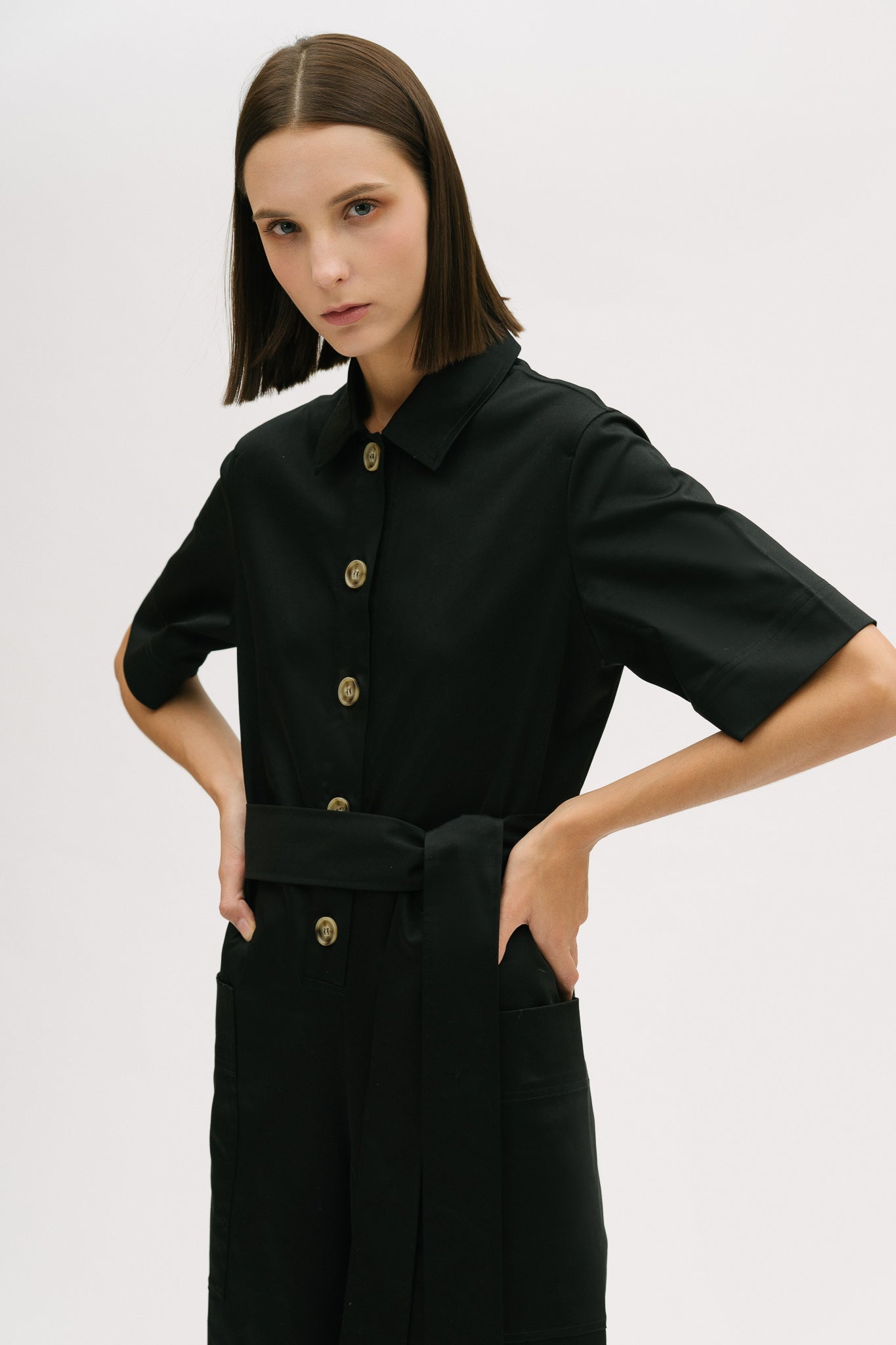 A woman with short brown hair is wearing a black collared jumpsuit with a tie belt.