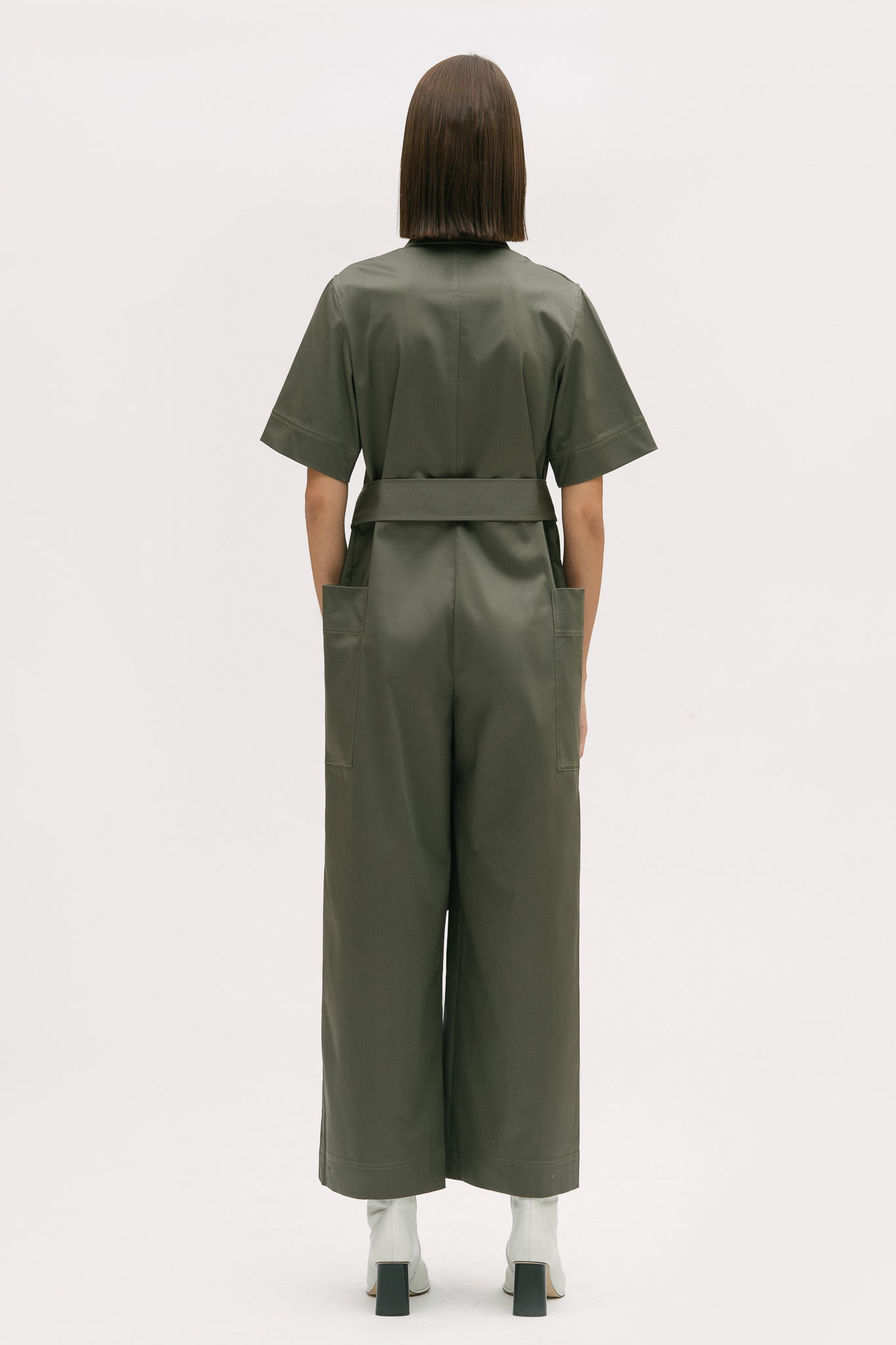 A woman with short brown hair is wearing a green collared jumpsuit with a tie belt. She is wearing white boots.