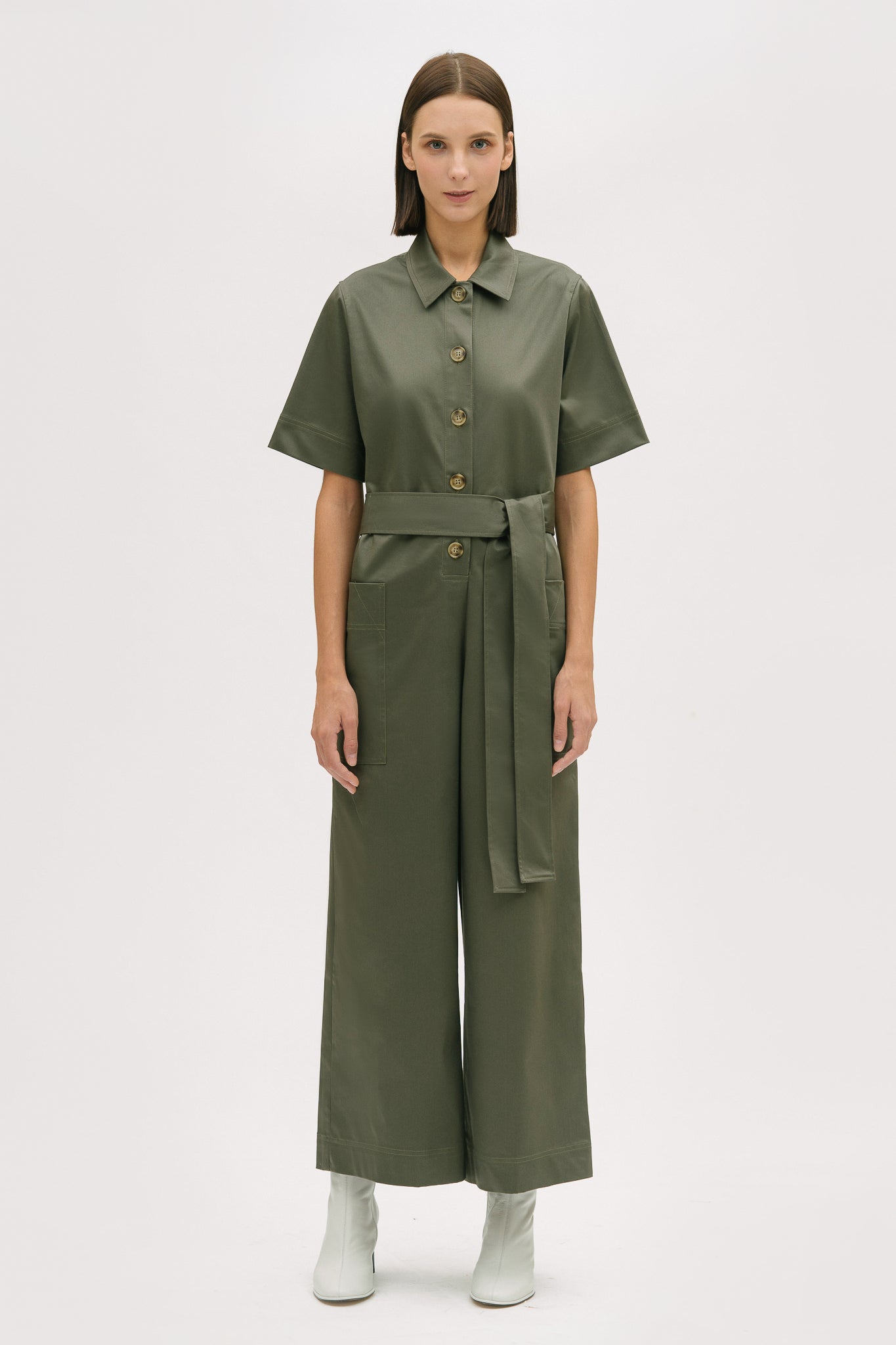 A woman with short brown hair is wearing a green collared jumpsuit with a tie belt. She is wearing white boots.