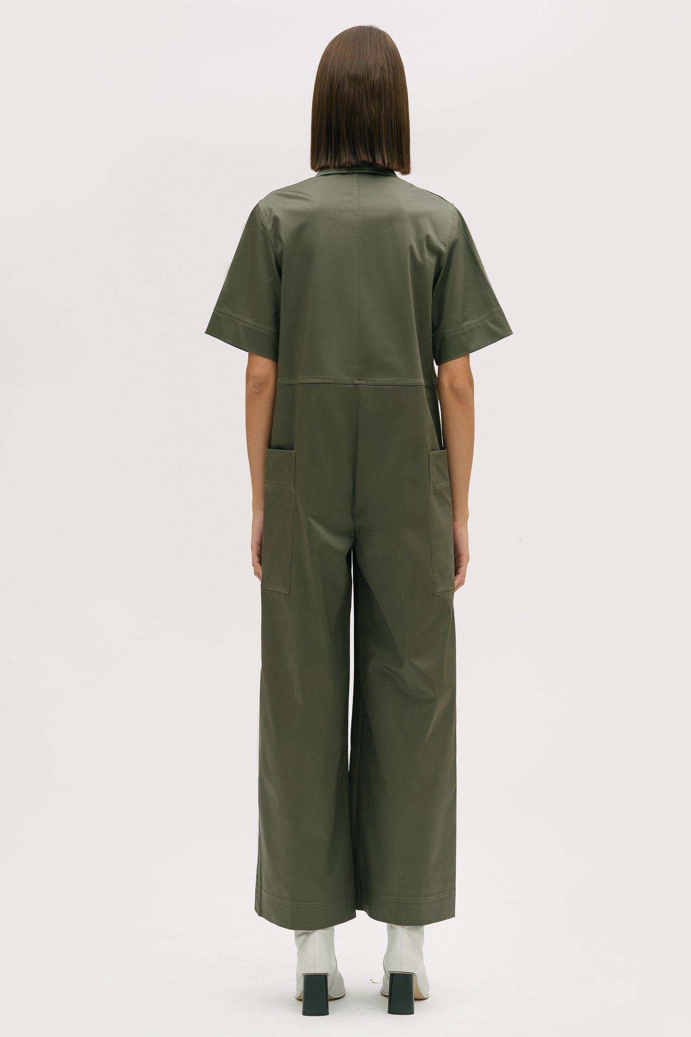 A woman with short brown hair is wearing a green collared jumpsuit. She is wearing white boots.