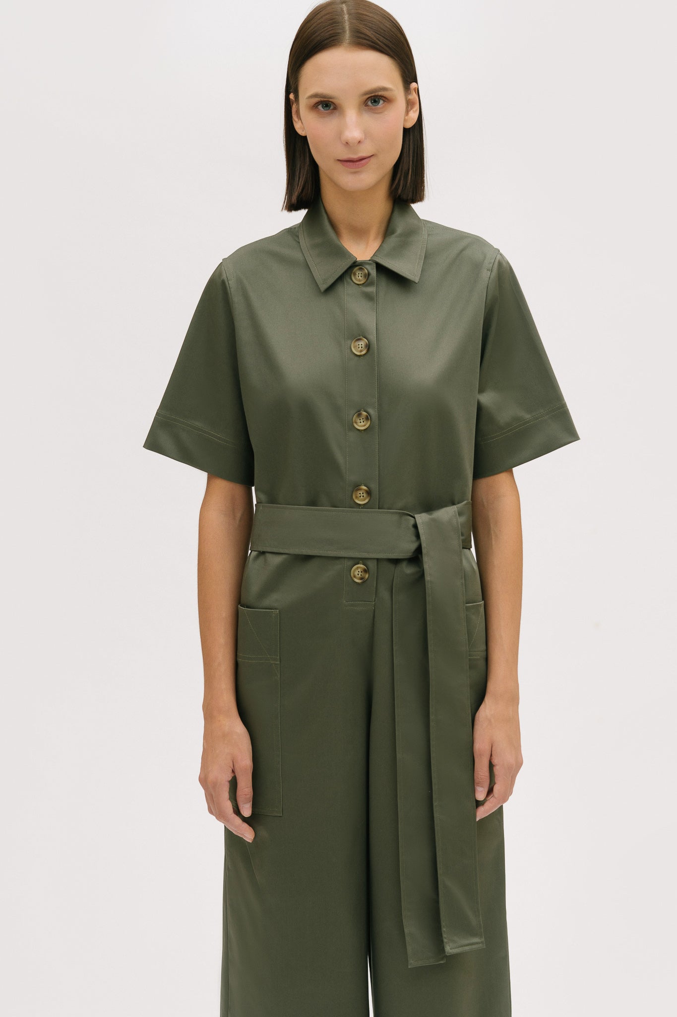A woman with short brown hair is wearing a green collared jumpsuit with a tie belt.