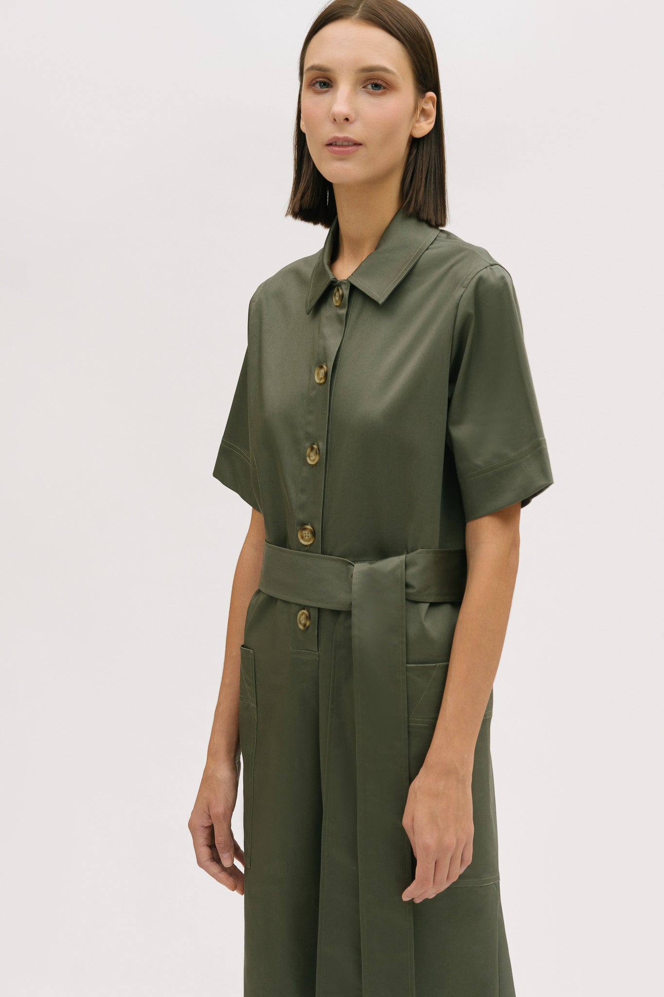 A woman with short brown hair is wearing a green collared jumpsuit with a tie belt.