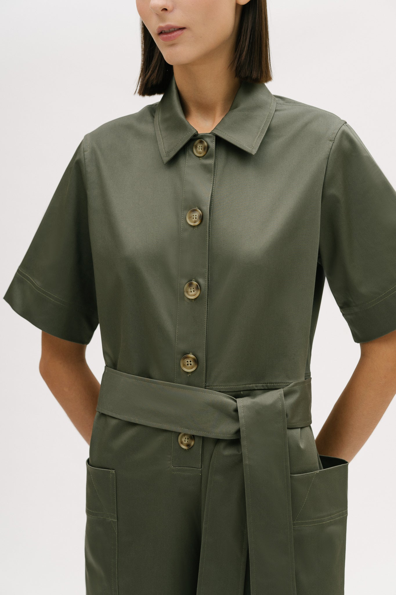 A woman with short brown hair is wearing a green collared jumpsuit with a tie belt.