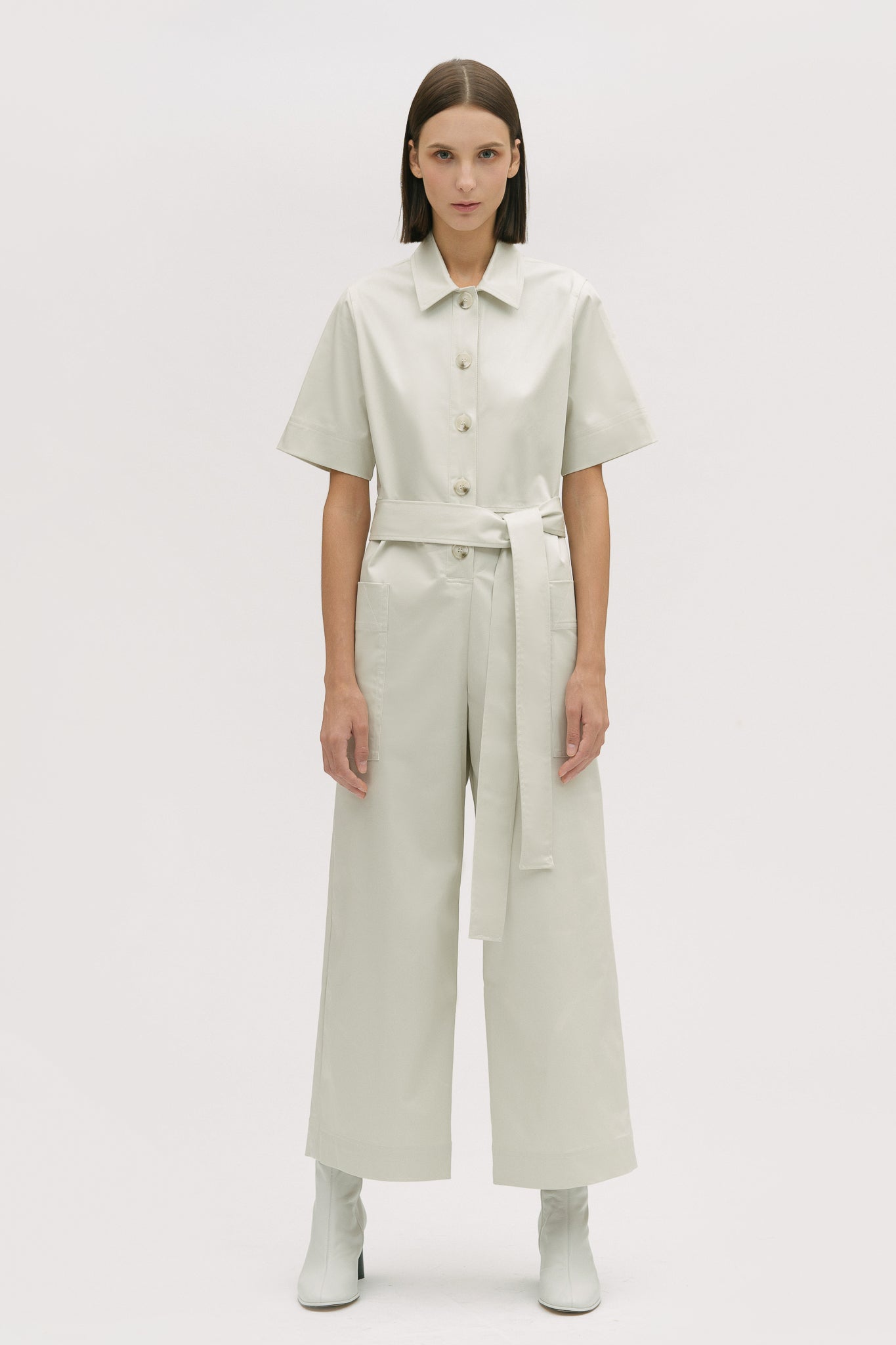 A woman with short brown hair is wearing a white collared jumpsuit with a tie belt. She is wearing white boots.