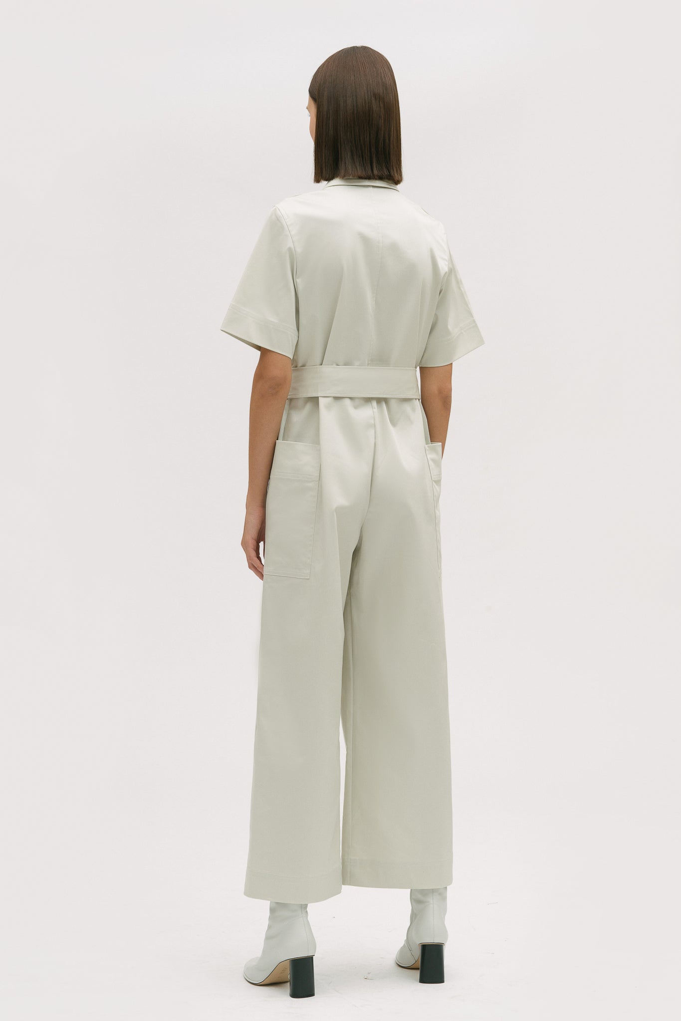 A woman with short brown hair is wearing a white collared jumpsuit with a tie belt. She is wearing white boots.