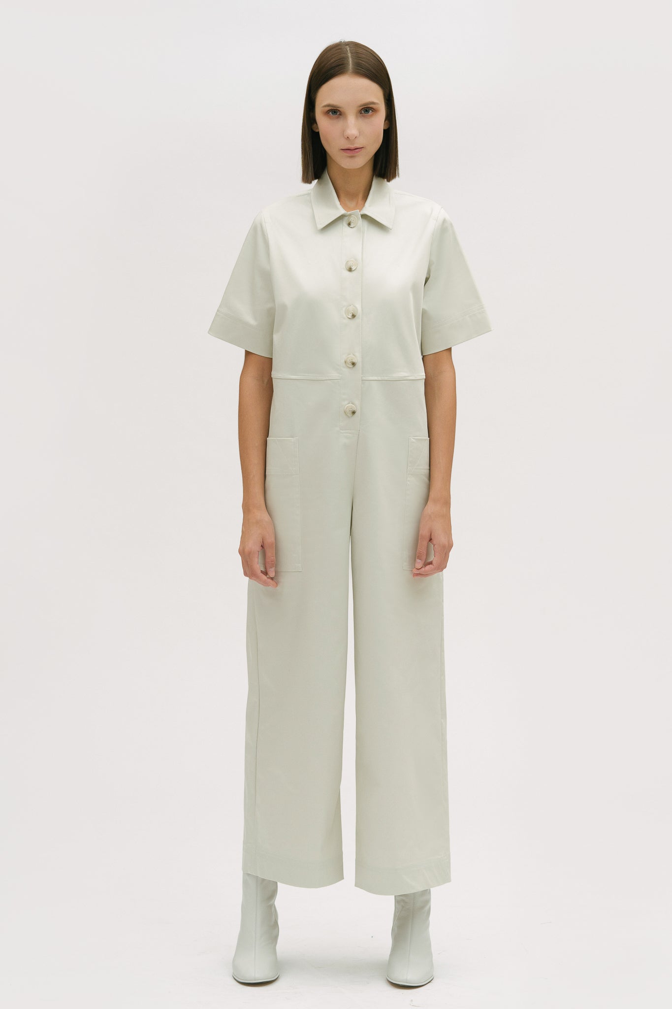 A woman with short brown hair is wearing a white collared jumpsuit. She is wearing white boots.
