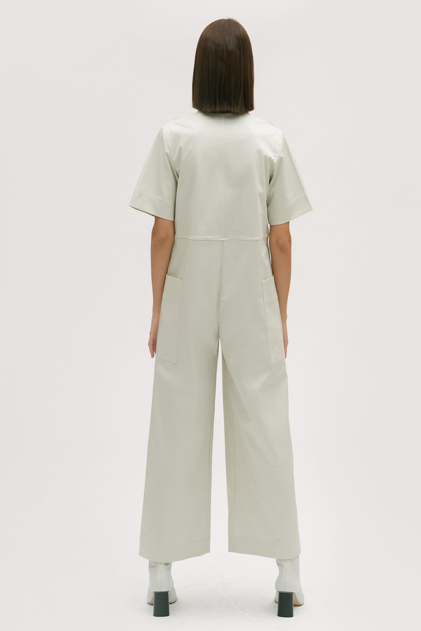 A woman with short brown hair is wearing a white collared jumpsuit. She is wearing white boots.