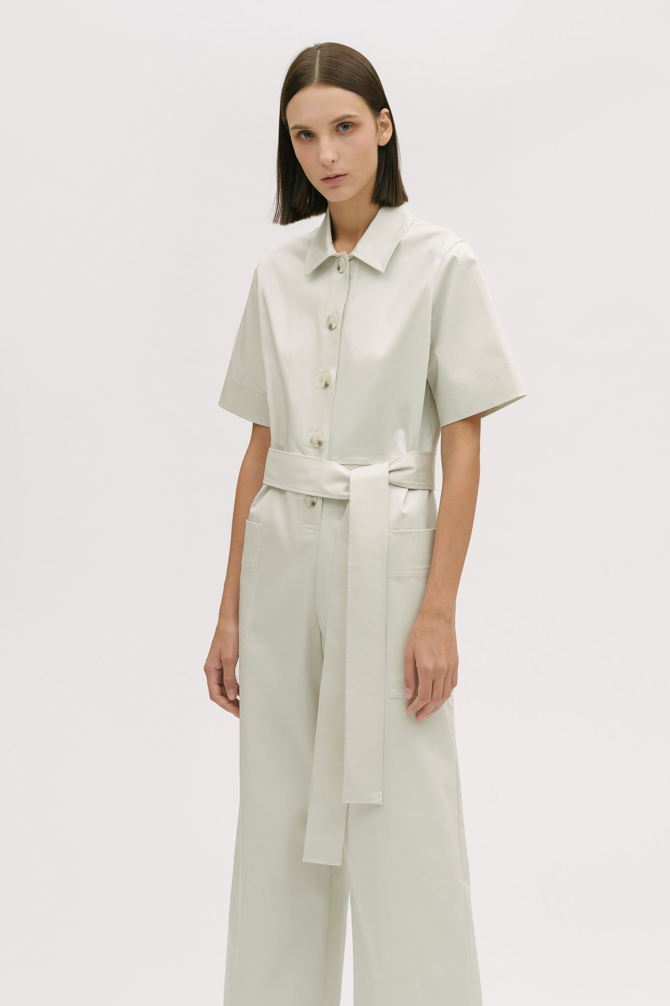 A woman with short brown hair is wearing a white collared jumpsuit with a tie belt.