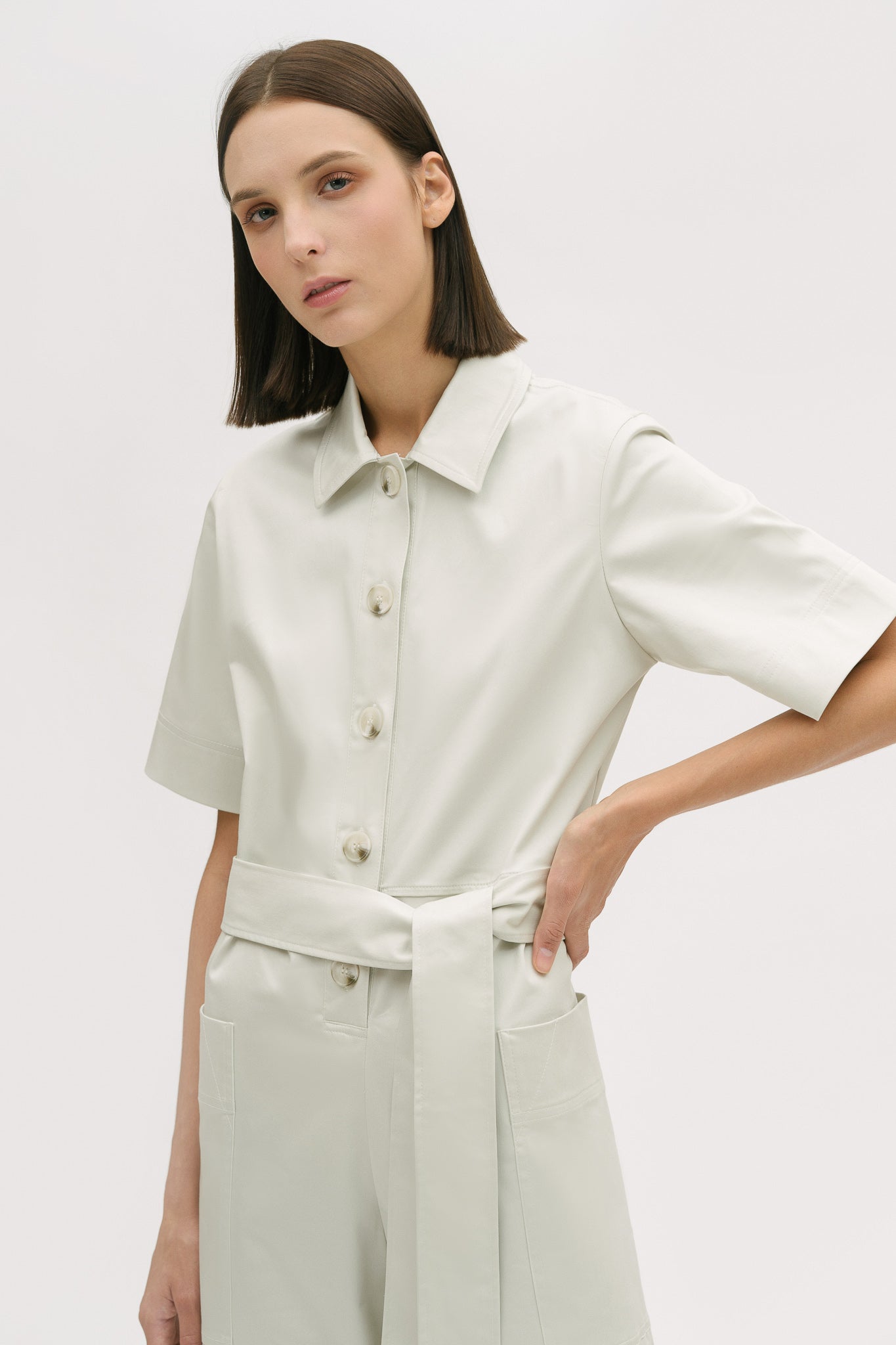 A woman with short brown hair is wearing a white collared jumpsuit with a tie belt.