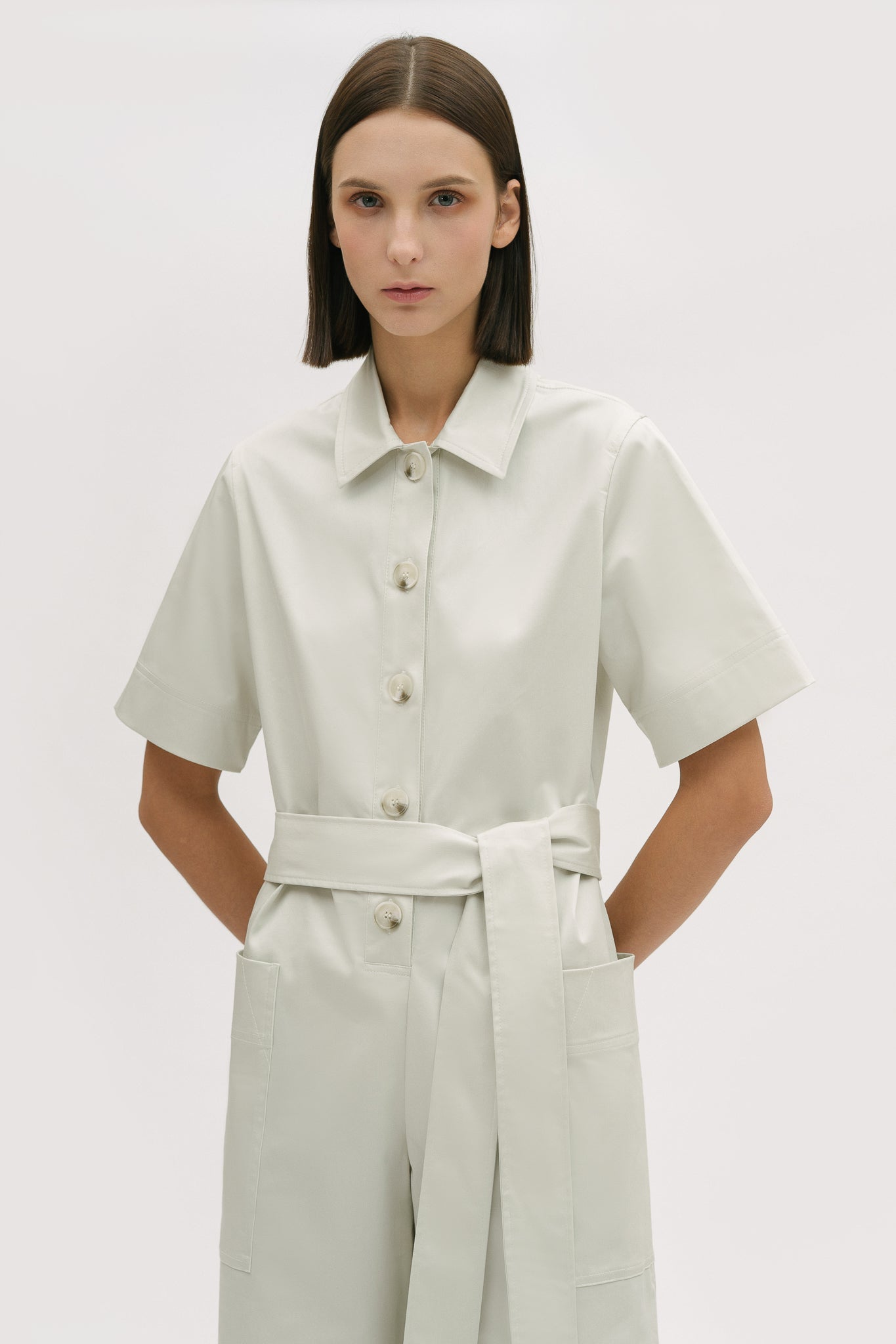 A woman with short brown hair is wearing a white collared jumpsuit with a tie belt. She is wearing white boots.